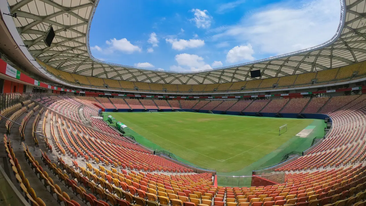 Campo e arquibancadas vazias da Arena da Amazônia, em Manaus, sob céu azul.