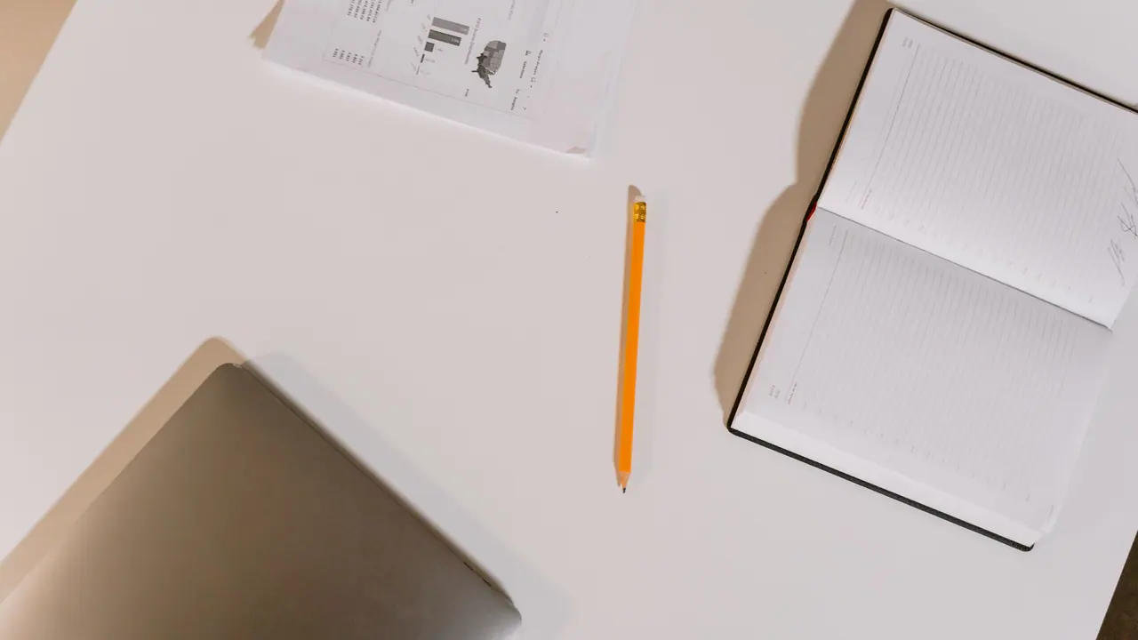 Caderno aberto, lápis e parte de um notebook sobre uma mesa clara.